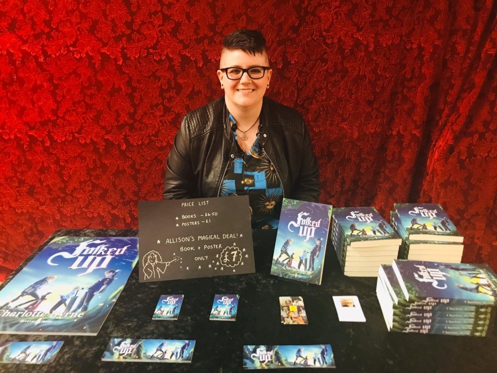 Charlotte Byrne sitting behind table with copies of Folked Up books and posters, and sign with prices on.
